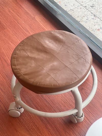 This is an overhead shot of a stool with a brown, quilted, leatherette seat and a light gray plastic base with wheels. The stool is positioned on a wooden floor with a reddish-brown hue. To the left of the stool, there's a dark gray metallic frame, likely part of a window or door, with a tiled floor visible beyond it. The tiles are a mix of white, gray, and speckled with dark flecks. The lighting suggests it is daytime, and the setting appears to be indoors, possibly in a home or commercial space in Gianyar, Indonesia. There is no discernible activity taking place, as the stool is stationary. A small, circular white sticker with some text is visible on the base of the stool.