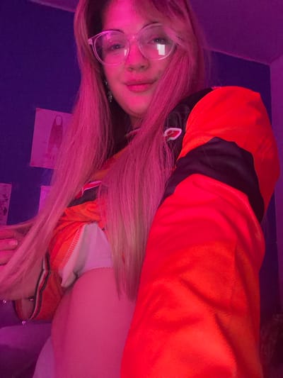 The image shows a young woman with pink hair, wearing glasses and an orange hoodie, lying on a purple surface. The location is not evident from the image itself and cannot be confirmed as being in Большая Шильна, Russia. There are some papers with writing on them behind her head.