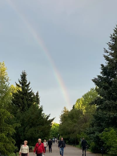 The image captures a vibrant rainbow arching across the sky above a tree-lined path in Subotica, Serbia. Several people are walking along the path, including one wearing a red jacket and another in a striped shirt. The scene suggests a park or green space within the city, with lush trees framing the sky and path. The overcast sky provides a backdrop for the colorful rainbow.