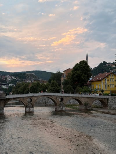This is a photograph of the Latin Bridge in Sarajevo, Bosnia and Herzegovina, taken at dusk. The bridge, constructed of stone, spans a shallow river with a rocky bed. Several people are visible walking across the bridge. In the background, buildings of various colors and architectural styles are clustered on a hillside, with trees interspersed among them. A tall, slender minaret of a mosque rises above the buildings. The sky above is filled with soft, scattered clouds, tinged with hues of orange and pink from the setting sun.