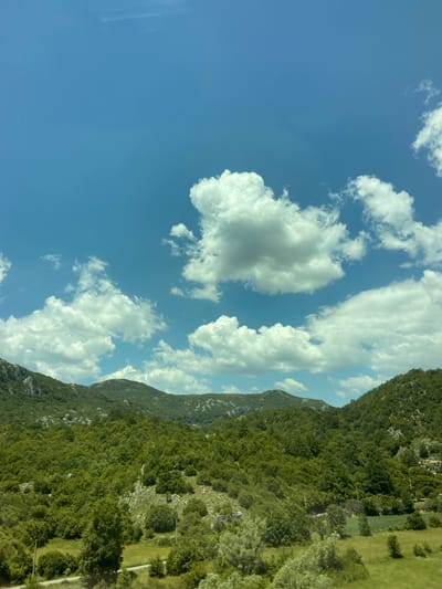 The image captures a scenic view of rolling green hills and mountains under a bright blue sky dotted with fluffy white clouds. The landscape is densely covered with lush green trees, with patches of lighter green meadows and some exposed rock formations visible on the slopes. In the foreground, there are some smaller trees and shrubs, and a glimpse of a road or path winding through the vegetation. In the distance, the peaks of the mountains fade into the horizon. A small cluster of buildings, suggesting a settlement, can be faintly seen on the far right side of the image, nestled among the trees on a hillside. This appears to be a rural, natural environment.