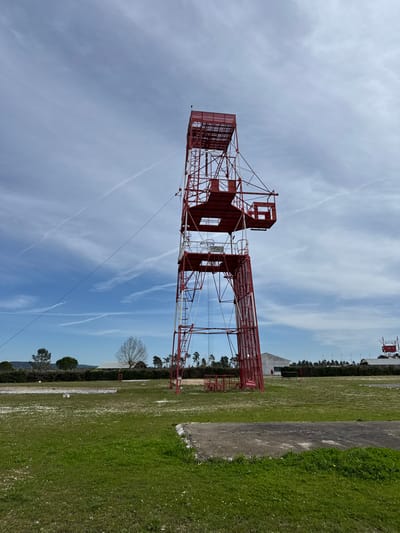 The image displays an outdoor scene in Praia do Ribatejo, Portugal, under a blue sky with scattered white clouds and multiple aircraft contrails. The primary subject is a tall, red-painted metal lattice tower, centrally positioned, extending vertically from a grassy field. This tower features multiple platforms and integrated ladders. A cage-like structure protrudes from its upper section, extending horizontally to the right. A taut cable extends diagonally downward from the upper left side of the tower, exiting the frame to the left. The ground is covered with green grass, interspersed with patches of small white flowers. A concrete slab partially covered by grass is visible in the lower right foreground. In the mid-ground, a line of green trees and bushes forms a border, with a leafless deciduous tree visible to the left. A light-colored building with a dark roofline is situated behind the central tower on the right side. Another, smaller red-painted metal structure, similar to the main tower, is visible in the far-right background. No individuals are present in the depicted scene.