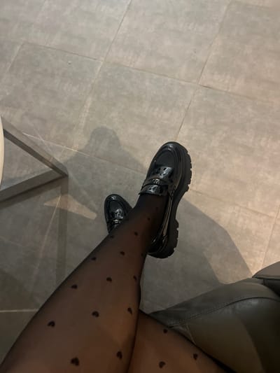 A person wearing black tights with small heart patterns is seated. The legs are crossed at the knees. The person is wearing black shiny penny loafers with thick soles. The background consists of gray floor tiles and a portion of a gray leather chair or couch to the right. The lighting casts shadows.