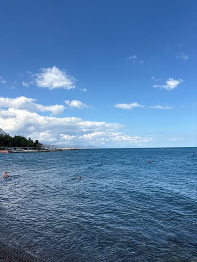 The image depicts a coastal scene in Alushta, Ukraine, on a clear day with a blue sky dotted with white clouds. The foreground features a pebbled beach transitioning into the rippling blue water of the sea. Two individuals are visible in the water, one closer to the shore and another further out, both appearing to be swimming or bathing. In the midground, a coastline is lined with lush green trees and buildings, including a large white structure that resembles a hotel or resort. A pier or promenade extends into the water, with several small boats or buoys visible. The horizon is defined by the distant outline of hills or mountains under the expansive sky. A single orange buoy is present in the water in the middle right portion of the frame.
