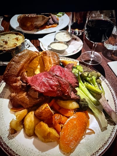 The image presents a close-up, overhead view of a meal served on a dark wooden table in what appears to be a dimly lit restaurant or pub. The primary focus is a large, ornate white plate, distinguished by a dark brown decorative rim, holding a substantial portion of food. Central to this plate is a generously sized, golden-brown Yorkshire pudding, partially filled with multiple slices of medium-rare roasted meat, identifiable by its reddish-pink interior and darker exterior crust. Alongside the meat and Yorkshire pudding are roasted golden-brown potato chunks, vibrant orange roasted root vegetables (likely carrots or parsnips), and a selection of green vegetables, including broccoli florets and elongated green stalks.
To the left of the main plate, a small, dark metal casserole dish or bowl contains a baked dish topped with a golden-brown crust, possibly a gratin or dauphinoise potatoes. In the background, another white plate holds a smaller serving of similar roasted meat and Yorkshire pudding. Two small, reflective metal ramekins, containing light-colored sauces, are positioned on a white saucer. To the right, a stemmed glass holds a dark red liquid, characteristic of red wine. Behind it, a tumbler contains a dark brown beverage, and a partially visible clear glass contains water. The table surface is dark wood, and a white napkin or paper is visible beneath the ramekins.
Based solely on the visual information contained within this image, there are no specific identifying features, such as landmarks, signage, or unique architectural elements, to determine its precise location within the city of Maidstone, United Kingdom.