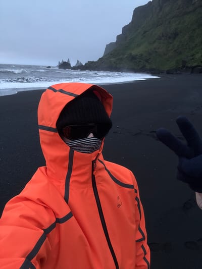 The image shows a person wearing an orange jacket, a black beanie, and gloves, standing on the black sand beach in Vik, Iceland. The person is making a peace sign with their gloved hand. The beach stretches out with waves crashing on the shore, and in the background, there are basalt columns. The sky is overcast.