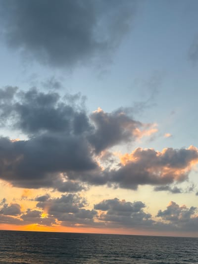 The image captures a seascape at dusk. The horizon is marked by a deep orange glow from the setting sun, which is partially obscured by dark, dramatic clouds. Rays of sunlight pierce through gaps in the clouds, illuminating the water below with a soft, golden light. The sea is a deep, textured blue-gray, with subtle ripples on its surface. Above, the sky transitions from the warm hues of sunset near the horizon to a pale blue, increasingly filled with scattered, dark clouds that are tinged with orange and pink where they catch the fading light. The scene appears to be a natural landscape. There are no people or man-made structures visible in the image.
