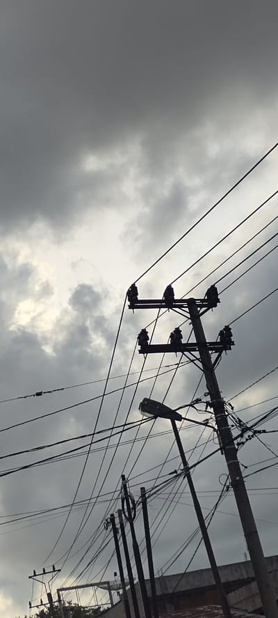 This image depicts a collection of utility poles and power lines against a dramatic, cloudy sky. The scene is likely from Keudegeudong, Indonesia, given the presence of dense overhead wiring typical of urban and semi-urban areas in the region.
In the foreground and mid-ground, several wooden utility poles stand tall, supporting a complex network of black power lines. One prominent pole has multiple crossbars with insulators, and on these insulators sit several silhouetted bird-like figures. These figures appear to be dark birds perched on the utility apparatus. Another pole, to the right, is also heavily laden with wires and features a street lamp mounted on its side. The street lamp itself is a dark, elongated fixture. In the lower left of the frame, a smaller utility pole with fewer lines and a few more silhouetted birds is visible, partially obscured by the dense wiring.
The background is dominated by a turbulent sky filled with dark, heavy clouds. Patches of lighter clouds suggest breaks in the cloud cover, allowing some illumination to filter through, creating a somewhat dramatic and overcast atmosphere. The overall impression is one of a utilitarian landscape set against a brooding sky, with the birds adding a touch of natural presence to the man-made infrastructure. There are no visible people or active interactions depicted.