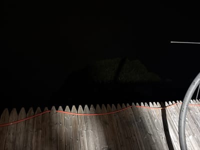 The image is a nighttime photograph taken in Riverhead, United States of America. In the foreground, a wooden fence with pointed pickets runs horizontally across the frame. An orange hose is draped over the top of the fence, following its contour. To the right of the fence, a curved, grey metal pipe is partially visible. The background is dark, with a faint silhouette of a tree or bush visible in the mid-ground, illuminated by an unseen light source. A thin, horizontal white line, possibly a light fixture or antenna, is visible in the upper right portion of the image against the dark sky.