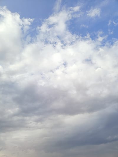 This is a full-frame, vertical, eye-level shot of a cloudy sky. The image is dominated by a variety of cloud formations, ranging from bright white, fluffy cumulus clouds at the top to darker, more ominous-looking nimbus clouds towards the bottom. Patches of blue sky are visible between the clouds, suggesting a partly cloudy day. The lighting suggests it could be daytime, possibly late afternoon or early evening given the darker tones of some of the clouds. There are no people, objects, or discernible location cues in the image. The focus is entirely on the atmospheric conditions of the sky. The image has a slightly grainy texture.