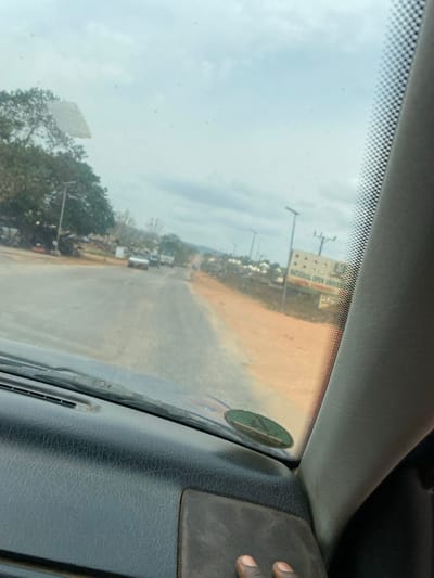 The image depicts a forward-facing view from inside a vehicle, capturing a road scene in Okengwe - Obehira, Nigeria. In the foreground, the vehicle's dark-colored dashboard is visible, with a section of the windshield above it. A dark-skinned human hand rests on the lower right portion of the dashboard. A green, oval sticker is affixed to the lower right of the windshield.
The road ahead is paved, appearing to be two lanes, extending into the distance under an overcast sky. The pavement exhibits patches of dirt and wear. A reddish-brown dirt shoulder borders the right side of the road. On the left side of the road, large green trees with dense foliage are prominent, with several dark-colored streetlights featuring solar panels standing along the roadside. Behind the trees, some indistinct structures are visible.
Multiple vehicles are present on the road in the mid-distance, including a light-colored sedan and a green truck, both facing away from the viewer. On the right side of the road, a large billboard or sign is visible, displaying "NATIONAL OPEN UNIVERSITY" in clear lettering, indicating a potential campus or study center in the vicinity. Beyond this sign and the dirt shoulder, various low-rise buildings are scattered. Distant hills or mountains are discernible on the horizon, beneath the light grey, cloudy sky. No direct human figures are clearly identifiable outside the vehicle.