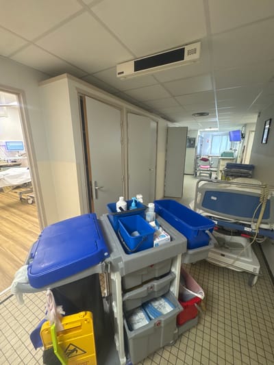 The image captures a hospital or clinical corridor, oriented vertically but depicting a horizontal view of the hallway. In the immediate foreground and to the right, a multi-tiered service cart, primarily gray with blue compartments, is parked on a beige tiled floor. The cart holds various supplies, including white bottles, blue containers, and white boxes labeled "polysans." A black trash bag is attached to the cart's lower level, next to a bright yellow "CAUTION WET FLOOR" sign featuring a falling person icon and a small green brush.
The corridor extends into the background, with white walls and a suspended ceiling composed of square panels. Several closed white doors line the left side of the hallway. A long, white rectangular device, possibly an air vent or heater, is mounted on the left wall. At the far end of the corridor, a window allows natural light into the space, and medical equipment such as beds, IV stands, and monitors are visible, suggesting patient rooms or treatment areas. One piece of medical equipment has a visible number "7" on it. No people are present, indicating a quiet moment in the facility. The bright lighting suggests it is daytime. The overall scene reflects a functional, maintained, and temporarily unoccupied medical environment.