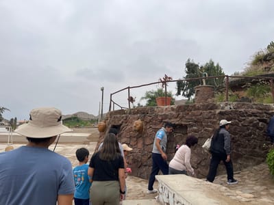 In Quilmaná, Peru, a group of people are ascending stone steps towards a rocky structure. The foreground features a person wearing a hat, another person with a dark shirt, and a child in a blue shirt. A stone wall with face sculptures and a wooden railing are visible. The environment includes trees, sky, and landscape in the distance.