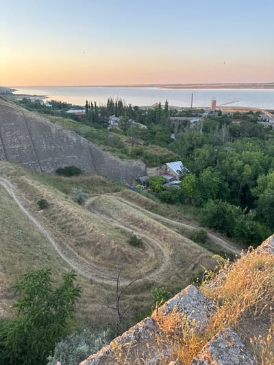 The image depicts a panoramic view overlooking a body of water and a settlement at sunset. In the foreground, a steep, rocky incline covered with dry grass and small purple flowers is visible. Below this, a winding dirt path cuts through a dry, grassy hillside. Further down, a cluster of trees and buildings, including a house with a white roof, are nestled amongst lush green foliage. Across the water, a distant shoreline is lined with buildings and industrial structures, including a tall tower with a light and a smaller tower with a red top. The sky displays a soft gradient of orange and blue, indicative of either sunrise or sunset. The scene appears to be the coastal area of Mizhlymanske, Ukraine.