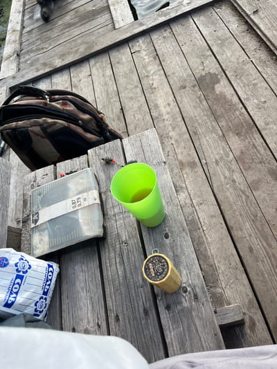 The image shows a collection of items resting on a weathered wooden surface, likely a dock or pier, given the presence of water visible in the bottom left corner. The scene appears to be outdoors, with the natural lighting suggesting it is daytime. The wooden planks are aged and have a rustic texture, with some dirt and debris visible.
The objects on the surface include a bright green plastic cup, a small cylindrical container with a label featuring ornate designs and text, a clear plastic tackle box containing fishing lures and supplies, and a patterned backpack. A fishing line with hooks and weights dangles from the green cup. A white plastic bag with blue lettering, likely containing salt, is also present in the lower right portion of the image.
The visible text on the cylindrical container includes "YONG ZHEN" and "YZH 242". The white bag displays the word "СОЛЬ" (salt) in large blue letters, with "«БЕЛАРУССКАЯ»" below it, indicating it is Belarusian salt. There are also some numbers on a white strip attached to the tackle box, including "55", "0.79", and "0.87".
The overall impression is of a scene set up for an outdoor activity, most likely fishing, given the tackle box, fishing line, and bait. The arrangement of items suggests they have been placed down temporarily. The lack of people or any indication of a specific event implies a moment captured during a casual outing. The setting of Вяззеўскі сельскі Савет, Belarus, places the scene in a rural Belarusian context.