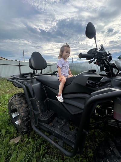 A young child is sitting on a black ATV, posed and looking to the side. The child is wearing a light purple t-shirt and white sandals. The ATV is parked on grass, with a metal fence and a cloudy sky in the background. The scene appears to be outdoors, possibly in a rural area given the grassy setting and fence. The lighting suggests it is daytime, with the sun partially obscured by clouds. The overall mood seems relaxed and casual, with the child appearing content. No visible text is present in the image. The provided location context is Kharnavy, Russia.