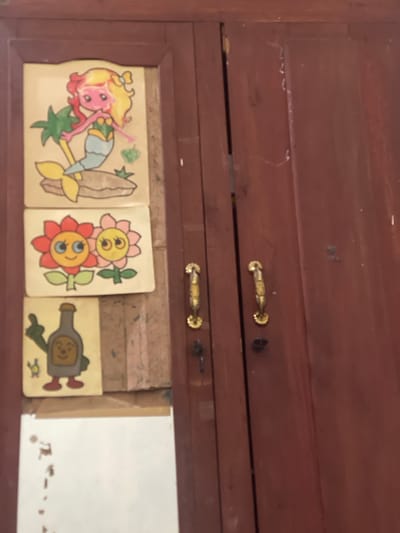 In the city of Anggaswangi, Indonesia, an aged wooden cabinet features three decorative decals affixed to its left door. The topmost decal depicts a cartoon mermaid with blonde and red hair, a green top, and a blue tail, positioned beside a palm tree and resting on a brown rock. Below the mermaid is a decal of two cartoon flowers, one red and one pink, each with wide blue eyes and smiling expressions. The bottom decal shows a cartoon bottle with arms and legs, wearing red shoes, giving a thumbs-up gesture, with a small blue and yellow creature to its left. The cabinet's right door is mostly visible, showing its dark red wooden surface and two ornate gold handles, one of which appears to have a key inserted. The overall environment suggests an indoor setting, possibly a home or shop, with the cabinet being a prominent fixture.