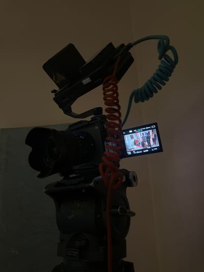 The image shows a close-up of a camera setup, likely for recording. A professional video camera, identified by the "alpha" logo and a lens with "G" and "FE 1.4/35" markings, is mounted on a Nest 590H tripod head. Attached to the camera's hot shoe is a small monitor displaying a scene with several people. A coiled orange cable connects to the monitor, and a blue coiled cable extends from the camera. The background is a plain, neutral wall, suggesting an indoor setting. The lighting is dim, casting long shadows and highlighting the equipment. There is no visible indication of the location being in Pakuro, Nigeria.