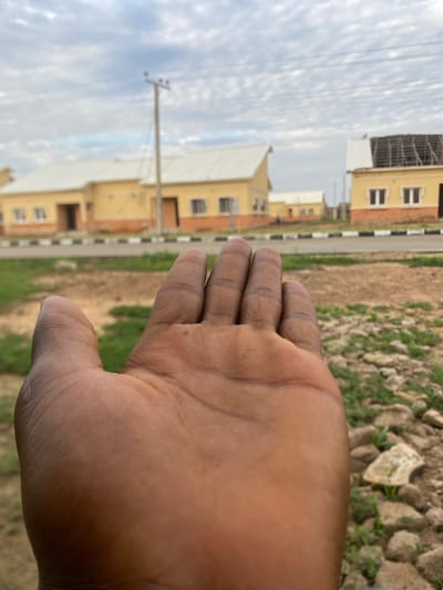 In Dumnga, Nigeria, an outdoor scene unfolds with a focus on a person's hand, palm up, in the foreground. The hand, exhibiting brown skin and visible lines, is positioned to occupy the lower left portion of the frame. Beyond the hand, the environment transitions to an unpaved area with scattered rocks and sparse greenery. Further in the background, a paved road with black and white markings runs horizontally across the image, separating the foreground from a row of yellow buildings. These structures appear to be residential or perhaps administrative buildings, featuring white roofs and numerous windows. A telephone pole stands near the buildings, and power lines stretch across the sky. The sky itself is overcast with a textured pattern of clouds. The overall impression is one of a developing area, with construction or recently completed structures in an outdoor, possibly semi-arid, setting.