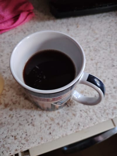 The image shows a close-up, high-angle shot of a white mug filled with dark liquid, likely coffee, resting on a speckled countertop. The mug has a blue and white design on its exterior and a blue stripe on its handle. To the upper left of the mug, a pink cloth is visible. The countertop has a speckled pattern of light and dark specks. The background is out of focus, but appears to be a kitchen or dining area. There are no people visible in the image. The scene is static and there is no indication of any action taking place. The lighting is adequate and appears to be from an overhead source. The image quality is good, with clear focus on the mug and its contents. The location is not specified in the image or its context.