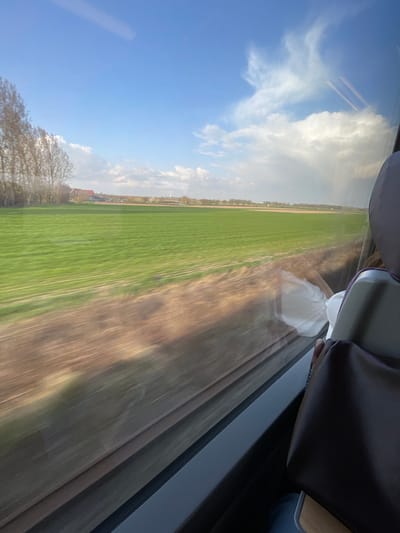 The image displays a rural landscape near Hochfelden, France, viewed from a passenger's perspective inside a moving train. The foreground outside the window exhibits motion blur, characterized by blurred brown earth and adjacent green agricultural fields displaying linear cultivation patterns. In the mid-ground, a cluster of buildings with red-tiled roofs is visible on the left, situated before a distant horizon marked by a sparse tree line. A tall, white, cylindrical structure, likely a water or communication tower, stands vertically on the far horizon. The sky is partially blue with scattered white cumulus clouds, becoming denser on the right. Inside the train, the upper portion of a dark brown passenger seatback with a grey headrest occupies the lower right quadrant. Reflections on the window glass include a crumpled white object and a human hand positioned near the window frame. No overt actions or interactions between individuals are discernible beyond the implicit presence of the person taking the photograph.