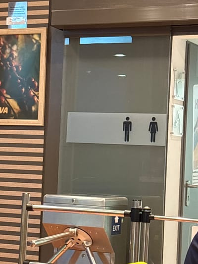 The image depicts a section of what appears to be a public restroom entrance. On the glass door, there are two restroom pictograms, one representing a male and the other a female. To the left of the restroom entrance, a turnstile mechanism is visible, with an "EXIT" sign and an upward-pointing arrow illuminated in blue on a dark blue background. A framed artwork depicting coffee beans hangs on a wall to the far left, adorned with horizontal wooden slats. The environment is indoors, with artificial lighting evident from the reflections on the glass. The city of Kalugerovo, Bulgaria, is not visually represented in the image, nor are there any discernible details that would indicate this specific location.