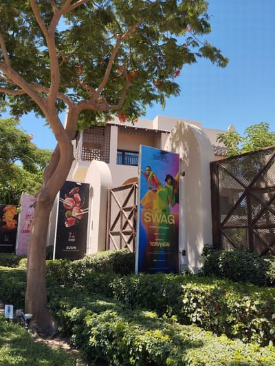 The image captures a sunlit outdoor scene featuring a building with a beige facade and arched doorways, framed by lush green foliage and the branches of a large tree with small red flowers. Several large vertical banners are displayed in front of the building. One banner, prominently featuring the word "SWAG" in large, colorful letters, advertises an event called "Elements" every Saturday, with music genres R&B and Hip Hop, and a resident DJ named ROMANIOS. Another banner displays a close-up image of sushi. Further to the left, another banner shows flames, likely advertising "Teppanyaki." The environment suggests a resort or entertainment venue, possibly a restaurant or club, located in Hurghada, Egypt, given the architectural style and the surrounding greenery. The sky is clear blue.