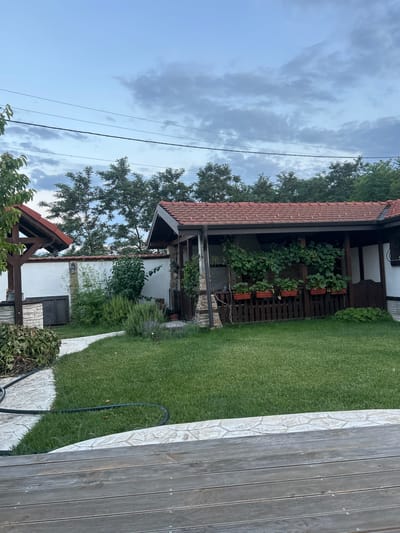 A slightly overcast sky hangs over a residential property in Voyvodino, Bulgaria. The scene depicts a backyard with a well-maintained lawn, a stone pathway, and a covered patio area. The patio, with its red tile roof and wooden railings, is adorned with potted plants and climbing vines. Adjacent to the patio is a white-walled structure with a smaller covered area and a wooden support beam. Trees line the background, and power lines are visible against the sky. A wooden deck occupies the foreground, suggesting a seating or lounging area. The overall impression is one of a peaceful, lived-in outdoor space.