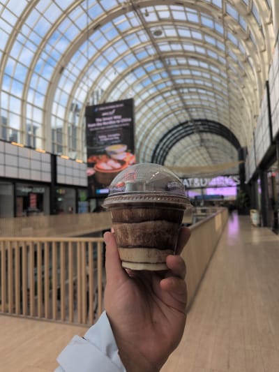 A person's hand holds a clear plastic cup containing a layered dessert. The dessert appears to have at least three distinct layers: a dark brown layer on top, a lighter tan layer in the middle, and a white layer at the bottom. The cup is covered with a clear plastic lid, and a thumb is visible on the right side of the hand, holding the cup.

The background suggests an indoor public space, likely a shopping center or market hall. A large, arched, glass-paned ceiling dominates the upper portion of the image, with sunlight filtering through the panes, casting a grid pattern on the interior. Below the ceiling, a large digital billboard displays an advertisement, partially obscured by the dessert cup. Further back, a wooden railing lines a walkway or balcony, separating it from the area below. Shops with visible signage are situated along the sides of the space. In the distance, illuminated text appears to read "EAT & DRINKS" above a brightly lit, purple-toned corridor.

The architectural style and the presence of a large modern atrium with a glass roof are characteristic of the **Kappeli** building, a historic pavilion located in Esplanadi Park in Helsinki, Finland. While the specific shops and the digital billboard are not identifiable as being exclusively within Kappeli itself, the overall ambiance of a spacious, light-filled indoor market or shopping area with a prominent atrium strongly aligns with the interior design of buildings like Kappeli, especially when considering modern renovations or adjacent structures that are part of the same urban development.