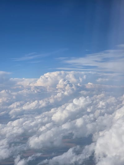 The image is an aerial view from an airplane, looking down at a vast expanse of clouds. The sky above is a clear, deep blue, transitioning into lighter shades as it meets the top layer of clouds. The clouds themselves are varied, with fluffy, cumulus formations dominating the scene. Some of these clouds are large and towering, while others are smaller and more dispersed. Below the main cloud layer, patches of land are visible, appearing as a muted tapestry of greens and browns. These landmasses suggest a rural or agricultural landscape. The lighting indicates it is daytime, with sunlight illuminating the tops of the clouds and casting shadows within their depths. The overall impression is one of immense scale and natural beauty, typical of an elevated perspective above the earth.