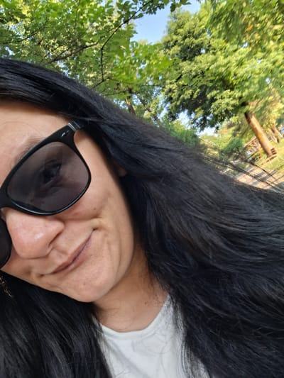 A woman with long, black hair and sunglasses is taking a selfie outdoors. She is smiling slightly, and her head is tilted to her left. The background shows lush green trees and a clear blue sky, suggesting a sunny day in a park or natural setting. The specific location in Gotse Delchev, Bulgaria, is not identifiable from the visual cues in the image.