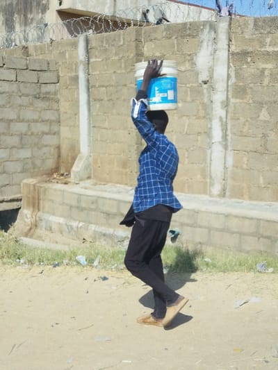 An outdoor scene in Babban Giji, Nigeria, depicts a lone individual walking from left to right on an unpaved dirt path. The person, viewed in profile, is wearing a blue and white plaid long-sleeved shirt, a dark undershirt, black trousers, and light brown slip-on shoes. They are actively balancing a white bucket, featuring a blue label, on their head, with their right hand raised to provide support. In the background, a light-colored concrete block wall runs horizontally across the frame, topped with razor wire. Below the wall, a low concrete structure, possibly a drainage channel, is visible. The ground is dry earth with sparse green vegetation and scattered debris. A clear, bright sky is visible in the upper portion of the background.
