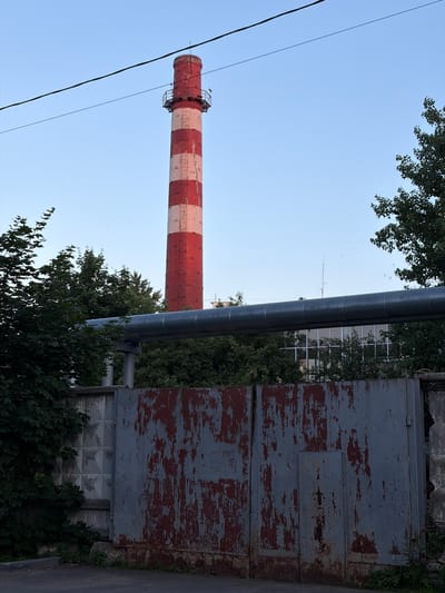 A tall, striped industrial chimney stands against a clear blue sky. The chimney is painted with alternating bands of red and white. A network of pipes runs horizontally in front of the chimney. To the right of the chimney is a dilapidated metal structure with peeling red and gray paint, possibly a large door or gate. Green trees and foliage surround the base of the structures. A power line runs diagonally through the frame from the bottom left towards the top. The scene appears to be outdoors on a clear day. There is no visible text.