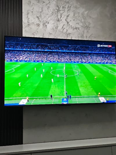 This image depicts an indoor domestic environment, purportedly located in Opaka, Bulgaria. A large, wall-mounted flat-screen television is centrally positioned, displaying a live football (soccer) match. The television screen shows a brightly lit green football pitch with white line markings and visible mowing stripes. Approximately two dozen football players are visible on the field; most wear white uniforms, while a smaller group near the midfield line wears dark-colored uniforms. Several officials, distinguishable by their yellow kits, are also present on the pitch. The stadium stands behind the field are filled with a dense crowd of spectators, appearing as a blurred mass. On-screen graphics in the top right corner display "b ACTION LIVE," with "UEFA CHAMPIONS LEAGUE" visible below. A blue advertising banner with "UEFA CHAMPIONS LEAGUE" branding is visible along the pitch's perimeter at the bottom of the screen. The television is mounted against a wall that features a dark, vertically ribbed panel on its left side and a light grey, textured, or marbled finish above and to its right. A low, white, minimalist shelf or cabinet unit is situated beneath the television.