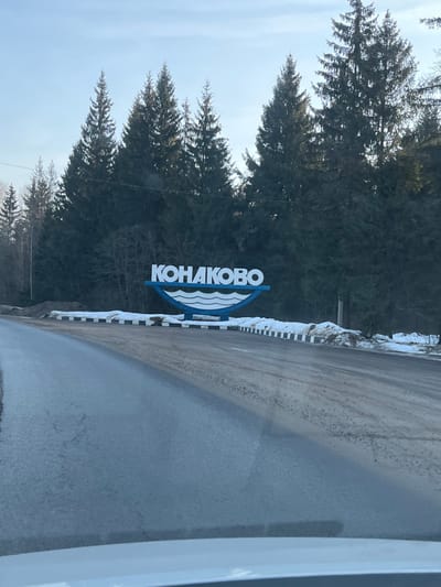 The image depicts the entrance to Konakovo, Russia, captured from the interior of a vehicle. A large, blue and white sign displaying the Cyrillic text "КОНАКОВО" (Konakovo) with a stylized wave motif is positioned on the right side of an asphalt road. The road surface shows patches of dirt and minor debris. Along the right shoulder, a low concrete barrier with alternating black and white stripes runs in front of the sign. Accumulations of dirty snow are present on the ground behind the barrier and around the sign's base. A dense forest composed primarily of tall, dark evergreen trees forms a backdrop behind the sign and extends across the upper half of the frame. The sky above the treeline is light blue and clear. No individuals or other vehicles are visible in the scene.