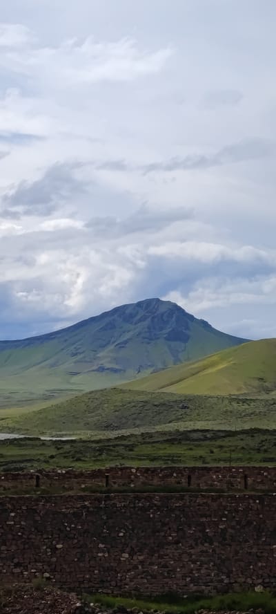 The image shows a landscape with a large, green mountain in the background. The sky is filled with white and gray clouds. In the midground, there are rolling green hills and a flat, grassy area. In the foreground, there is a dark, rocky wall structure. The location is likely near Dashtadem, Armenia, based on the style of the stone wall and overall landscape. There are no people visible in the image.