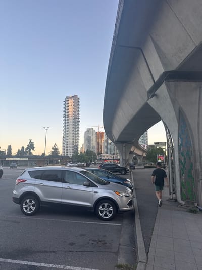 The image shows a parking lot adjacent to a pedestrian walkway and a concrete overpass, likely part of a SkyTrain line in Coquitlam, Canada. The setting is urban, with several tall buildings visible in the background, including a prominent, modern skyscraper reflecting the golden light of what appears to be late afternoon or early evening. A single man is walking away from the camera on the right side of the image, wearing a black t-shirt and green shorts. In the foreground, a silver SUV is parked, and other vehicles are present in the lot. The concrete structure of the overpass dominates the upper right portion of the frame, showcasing its architectural design. The sky is clear and pale blue.