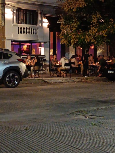 The image depicts a restaurant in Montevideo, Uruguay, with outdoor seating. Several people are seated at tables along the sidewalk, appearing to be dining or socializing. A car is parked on the street next to the tables. The restaurant building is two stories tall, with a balcony on the upper level. The street is paved and appears to be in an urban setting. The scene is at night, with light emanating from the restaurant and streetlights.