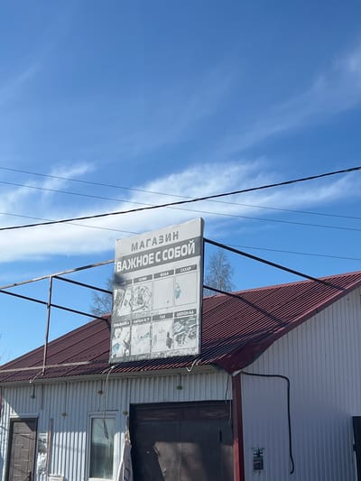 The photograph shows a building with a sign that reads "МАГАЗИН ВАЖНОЕ С СОБОЙ" (translated to "SHOP IMPORTANT WITH YOU"), with further details about items for sale below, including tea, coffee, water, sugar, windshield wiper fluid, noodles, gas, and chocolate. The building is a one-story structure with a metal roof and corrugated metal siding. There is a door and a window visible on the side of the building. Power lines run across the top left of the frame, and the sky is blue with thin clouds. The image appears to be taken in Gory, Russia, based on the language of the signage.