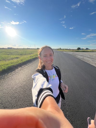 A woman is taking a selfie outdoors on a sunny day. She is wearing a white t-shirt with colorful graphics and a black and white striped jacket. The background shows a paved road with a grassy field on the left and a clear blue sky with some scattered clouds on the right. The sun is shining brightly, casting a glare in the sky. The scene appears to be in a rural or open area, possibly an airfield or a long road in the countryside. The woman is smiling, suggesting a cheerful mood. The time of day is likely late afternoon or early evening, given the low angle of the sun. The location context is Pivkino, Russia. There is some visible text on the woman's t-shirt that appears to be "GIRL" and some fruit graphics.