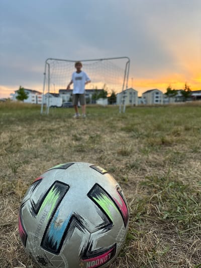 A soccer ball is in the foreground of a grassy field. In the background, a young person stands in front of a soccer goal. The sun is setting, casting a warm orange glow across the sky. In the distance, houses and trees are visible, suggesting a suburban or semi-rural setting. This scene is located in Bönnigheim, Germany.