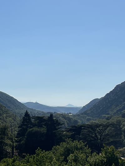 The image depicts a serene mountainous landscape under a clear blue sky, likely during the daytime. The composition is dominated by lush green hills and mountains, heavily forested with a variety of trees. A prominent mountain range with a distinct, rugged silhouette occupies the right side of the frame, fading into softer, layered ranges in the distance. The sky is a vast expanse of light blue, transitioning to a paler hue near the horizon, suggesting a bright, clear day.
In the mid-ground, a few man-made structures, possibly buildings or power lines, are visible nestled within the valleys and on the slopes of the distant mountains, hinting at human habitation within this natural setting. The density of the foliage suggests a temperate climate and a well-established forest ecosystem. The overall impression is one of natural beauty and tranquility. There is no visible text in the image.