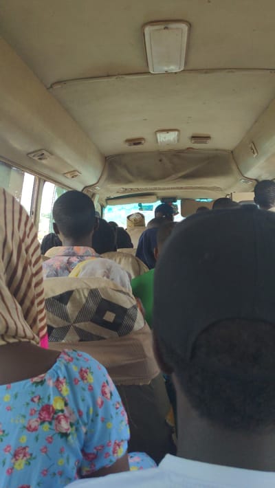 The image captures the interior of a public transport vehicle, likely a bus or large van, filled with passengers. The perspective is from behind individuals in the front seats, looking towards the front of the vehicle. The entire scene is oriented as if the camera was rotated 90 degrees clockwise.
Multiple subjects are visible, primarily from the back of their heads and shoulders, seated within the vehicle. In the immediate foreground, a person on the right wears a light blue top with a vibrant floral pattern of pink, yellow, and green flowers. Next to them, another passenger is seen wearing a garment with a distinct geometric pattern of black and off-white squares. Further ahead, several other passengers are seated, some wearing dark caps or hats. A person towards the front has a dark blue top, and a bright green garment is visible on another.
The interior of the vehicle features a light-colored, possibly off-white or cream, ceiling and upper wall lining that appears somewhat worn. Rectangular overhead fixtures, possibly lights or vents, are mounted on the ceiling. Through the front windows, a bright sky or exterior is visible, indicating it is daytime. The packed seating arrangement and vehicle type suggest a common public transportation scene in Kugbo, Nigeria. No discernible text is visible within the image. The activity depicted is simply people traveling or waiting inside the vehicle.