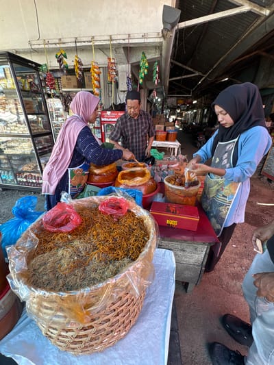 In the city of Grong-Grong, Indonesia, at an outdoor market stall, two women and one man are engaged in commerce. A woman wearing a pink hijab and a blue and white patterned shirt, with a blue apron featuring a teddy bear motif, stands behind a large woven basket filled with dried, thin noodles. To her right, a woman in a black hijab and a blue long-sleeved shirt, also wearing an apron, is packaging food items from a plastic tub into a plastic bag. A man in a plaid shirt and a black cap stands between them, overseeing the transactions. In front of the women are several containers and a red box labeled "BOSS." The stall is lined with various goods, including snacks and drinks displayed in a glass-fronted refrigerator. The market appears to be under a corrugated metal roof, suggesting an open-air or semi-enclosed environment, with motorcycles visible in the background. A person's legs and feet, wearing light blue jeans and black sneakers, are visible in the foreground, with their hand holding currency.