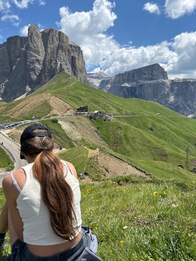 A young woman with long, reddish-brown hair pulled back into a ponytail wears a white tank top and a black baseball cap. She is seated in a grassy, sunlit meadow dotted with small yellow flowers, her back to the camera. In the background, a winding road curves up a lush green mountainside, with a parking lot filled with cars visible on the left. Several buildings, possibly restaurants or lodging, are situated along the road and on the slopes. Towering, rugged mountain peaks dominate the horizon under a bright blue sky scattered with fluffy white clouds. The landscape appears to be a mountain resort area in a remote, natural setting.
