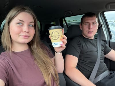 Inside a vehicle in Совхозный, Russia, a light-skinned female and a light-skinned male are seated. The female, positioned on the left, has long light brown hair, blue eyes, and visible freckles. She is wearing a dark mauve short-sleeved t-shirt and is holding a light brown paper coffee cup with a dark lid in her right hand. The cup features a "ГАЗПРОМ" logo and the Russian text "СВЕЖИЙ КОФЕ" (Fresh Coffee). Her fingernails are adorned with multi-colored polish. The male, seated on the right in the driver's seat, has short dark hair and blue eyes. He is wearing a black short-sleeved athletic shirt and a gray seatbelt across his chest and lap. Both individuals are looking towards the camera. The vehicle's interior appears dark, and through the windows, other vehicles and a cloudy sky are visible, indicating an outdoor setting.