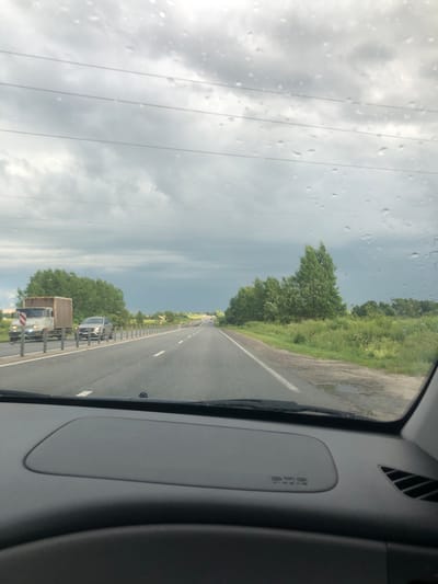 A POV shot from inside a vehicle shows a highway under a cloudy sky. The windshield has raindrops on it. The road has multiple lanes and is bordered by a metal guardrail and vegetation. Several vehicles are visible on the road, including a white truck with a brown cargo box and a silver car. Trees and fields line the sides of the road, and power lines are visible in the sky. The dashboard of the vehicle is visible in the foreground.