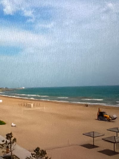 The image depicts a wide, sandy beach in Lopatino, Russia, with the turquoise sea and a pale blue sky dominating the upper half. In the distance, a white lighthouse or structure stands on a rocky outcrop. The beach itself is mostly empty, with gentle waves lapping the shore. A yellow construction vehicle with a front loader is visible on the sand, appearing to be in operation. Further down the beach, several umbrella-like structures are set up, casting shadows on the sand. In the foreground, a portion of a paved area with some trees is visible, suggesting a walkway or promenade adjacent to the beach. The overall impression is one of a quiet, slightly overcast day at the seaside, with some light industrial activity taking place on the beach.