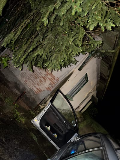 The image shows a nighttime scene in or near Umarevtsi, Bulgaria. A vehicle with an open door is parked alongside an old, possibly abandoned building. The building has weathered, light-colored walls, a dark window frame and a brick section near the ground. A large evergreen tree is situated to the left of the building. The environment appears damp, with vegetation visible on the ground near the car. The open car door contains trash. No people are visible in the frame.