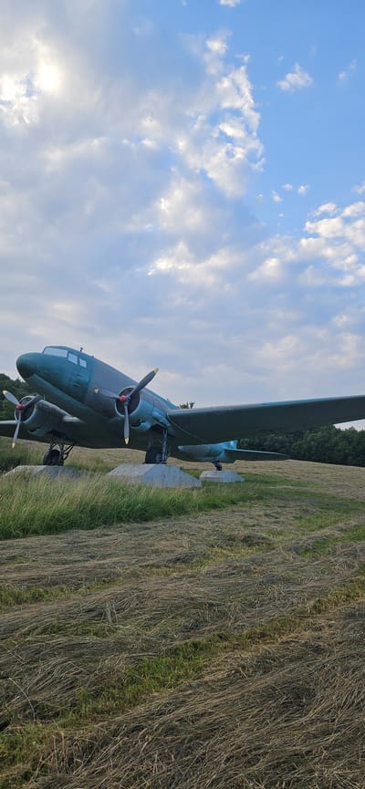 An old, teal-colored propeller airplane is parked on a grassy field in Vyšný Komárnik, Slovakia. The airplane is supported by concrete blocks and sits on landing gear. The sky above is blue with scattered white clouds, and the sun is partially visible. The field is covered in dry, matted grass, with some green grass peeking through. Trees are visible in the background. The scene appears to be a static display of the aircraft.